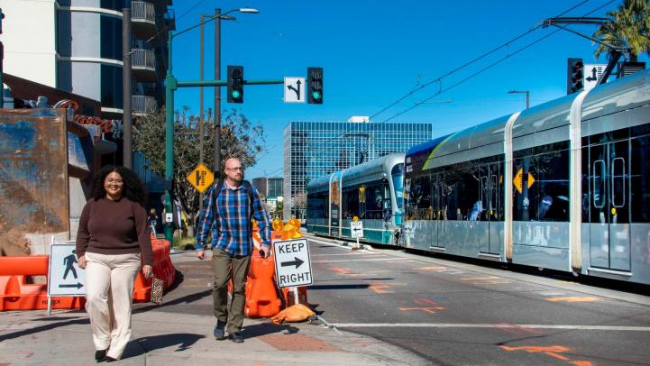 Two people walking through a construction zone next to VM light rail.