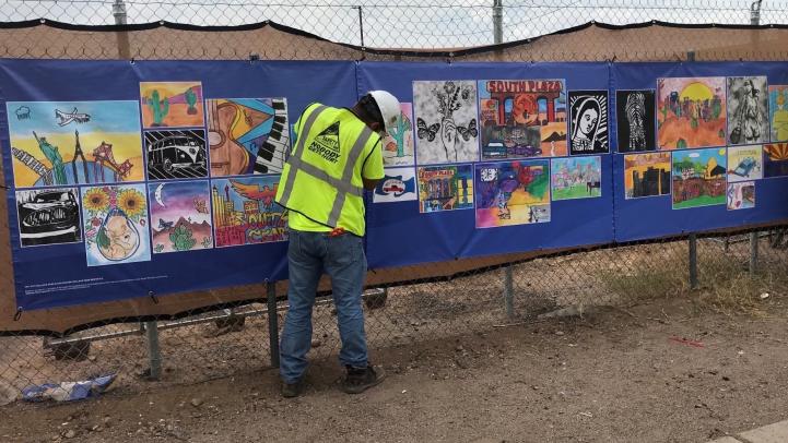 Construction worker hanging student artwork banner on fencing along Central Ave