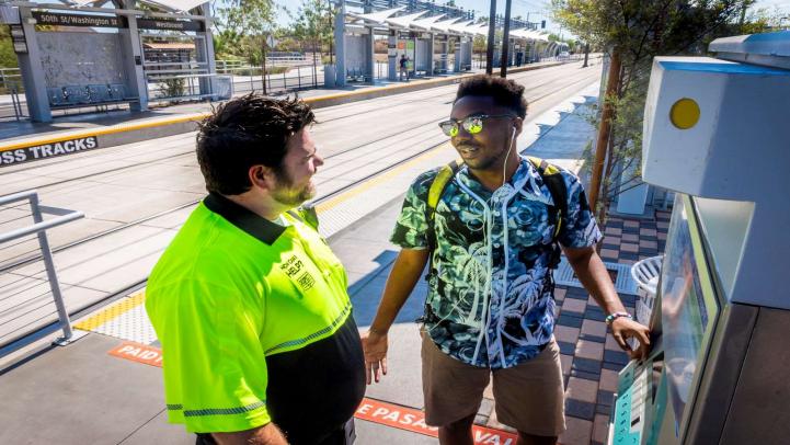 Customer Experience Coordinator assists rail passenger Coordinator in black and yellow uniform with male rider at farebox